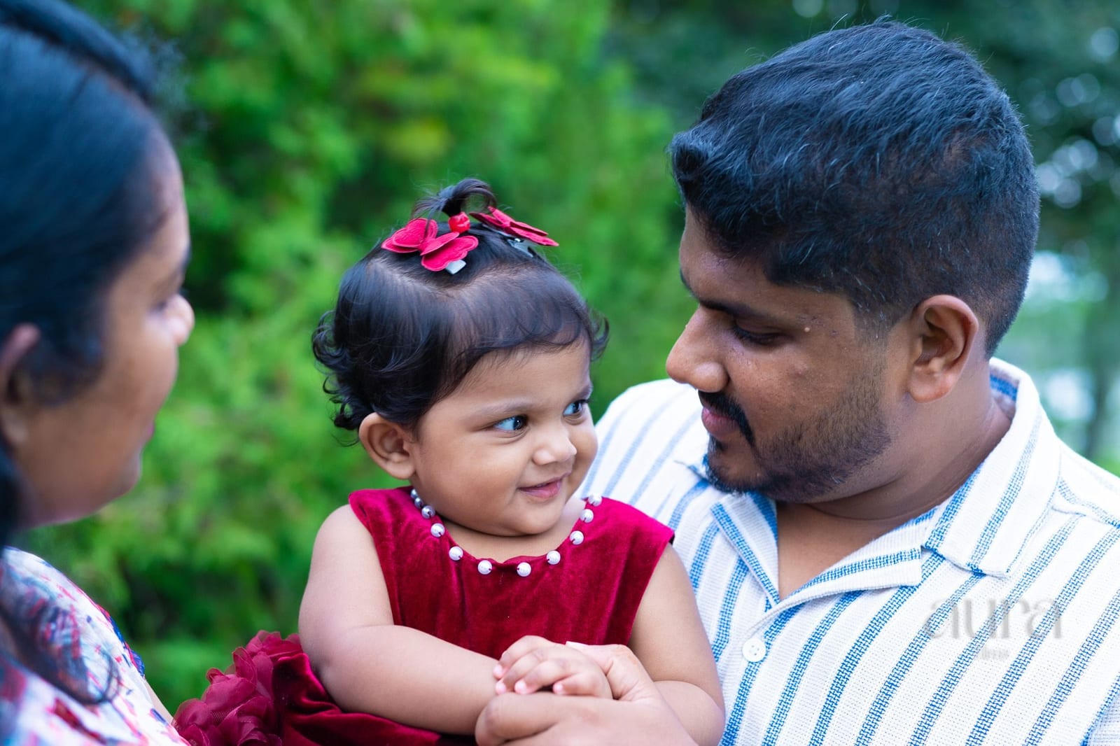 Parents lean in close to their toddler, who wears red bows in her hair