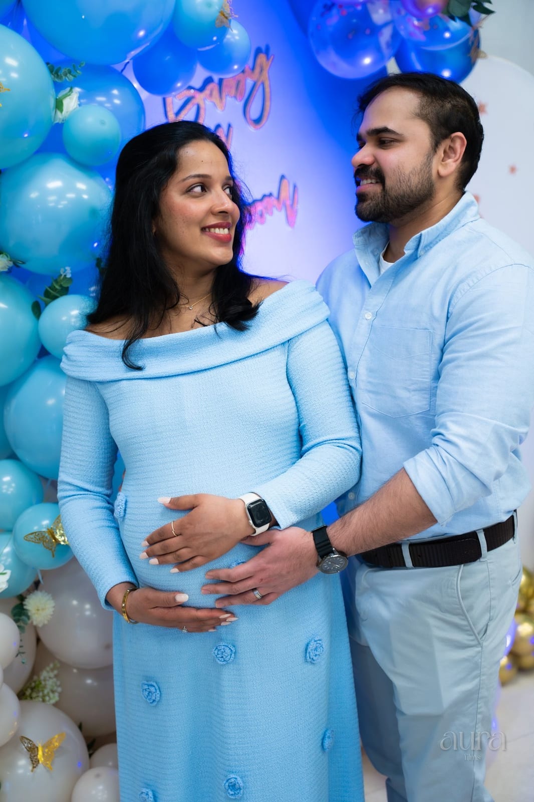An expecting couple smile together beside blue balloons