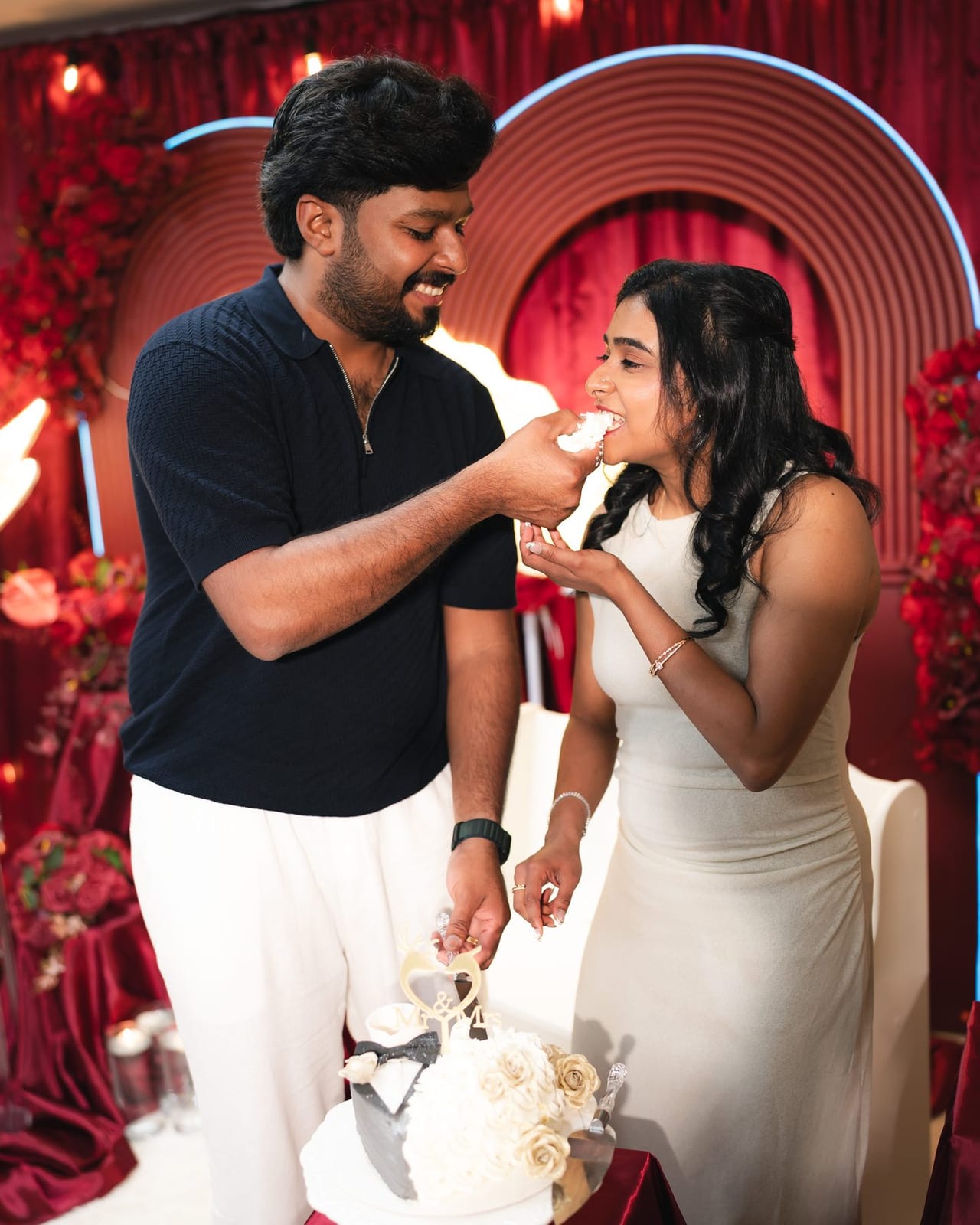 A couple feed each other cake in front of a red floral wall