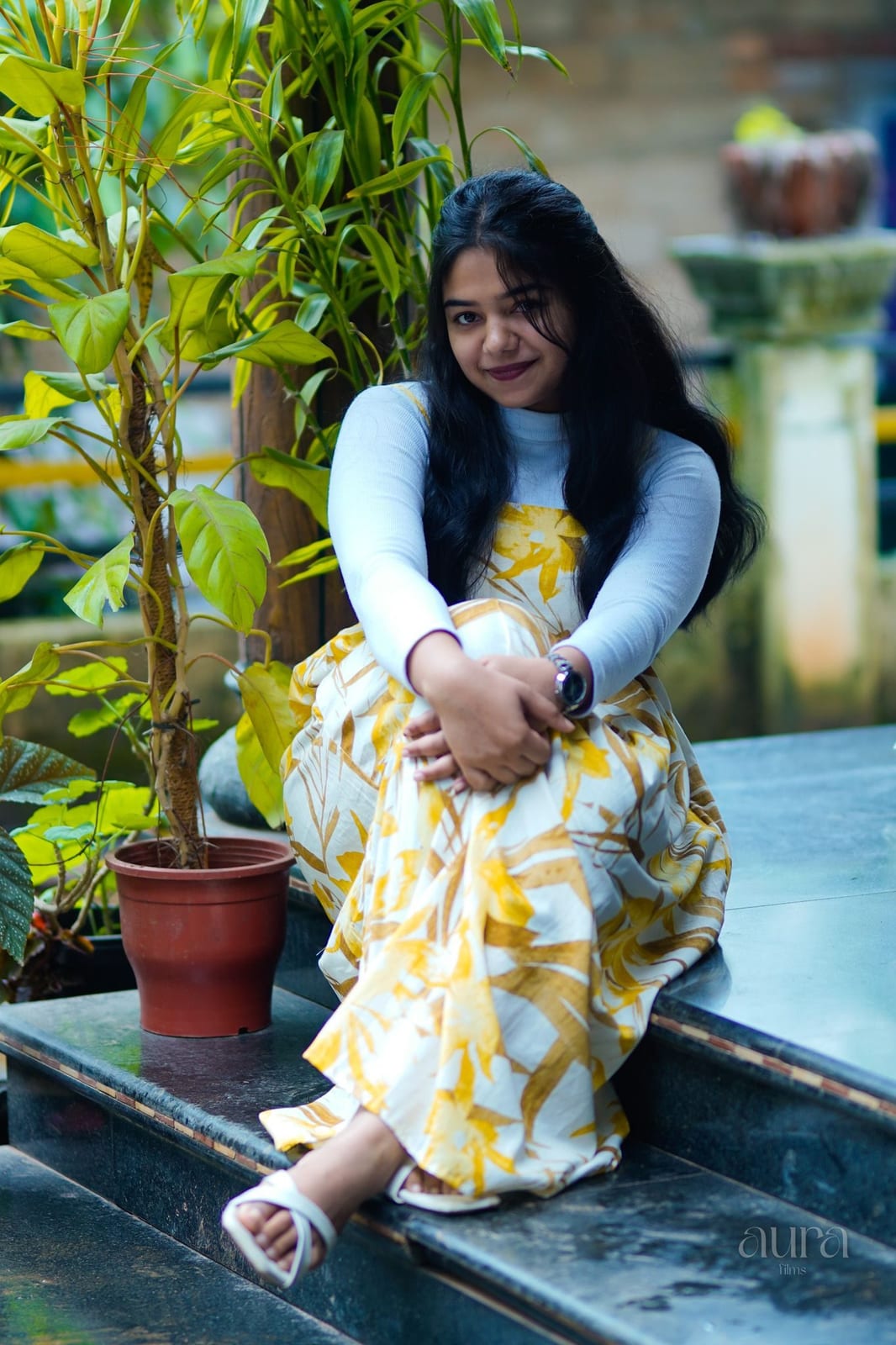 A woman in a yellow floral dress sits among potted plants