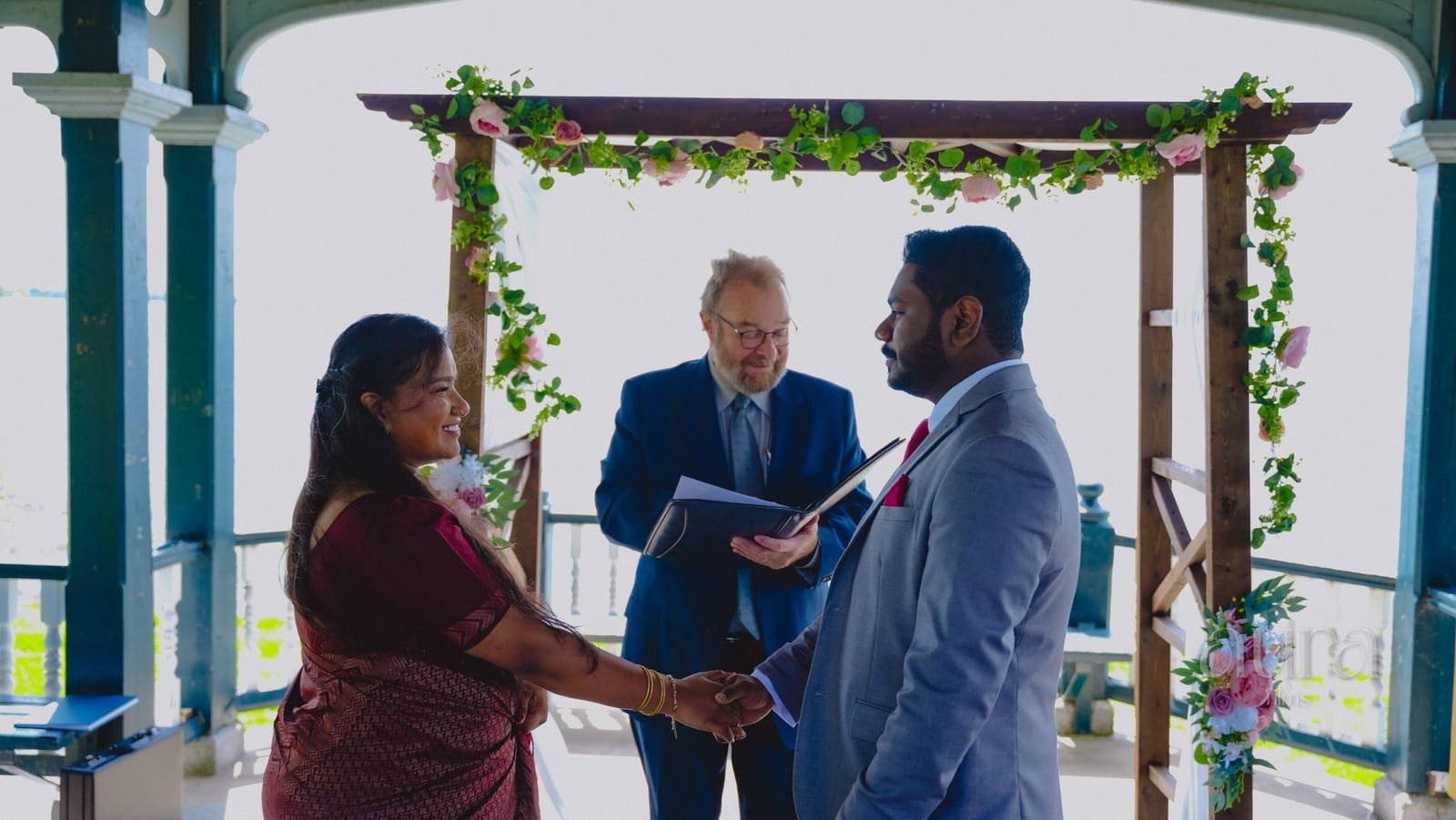 A couple exchange vows under a flower-covered arbour beside the lake